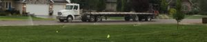 An empty flatbed semi truck used to transport the sod parked on the road. In the foreground is a sod lawn being watered.